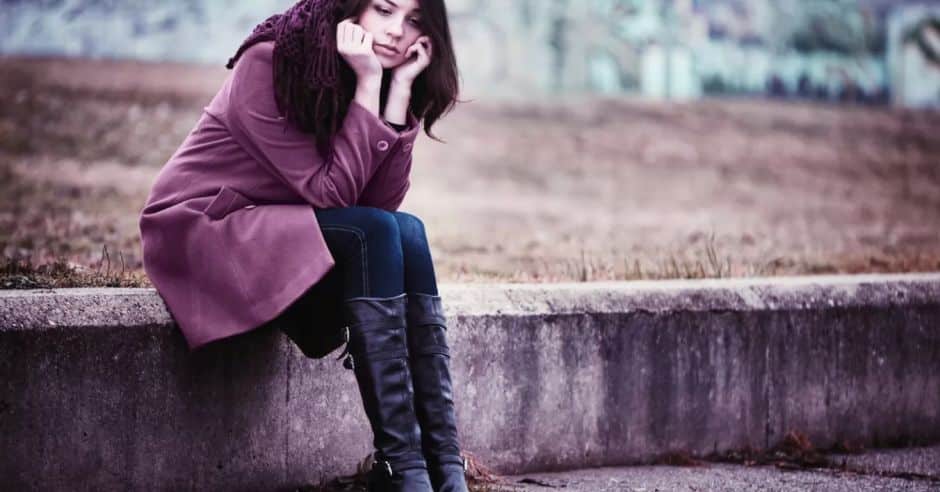 A young woman with dark hair, wearing a purple coat and scarf, sits alone on a concrete ledge outdoors, looking down with a somber expression, conveying a mood of loneliness and contemplation in self-pity