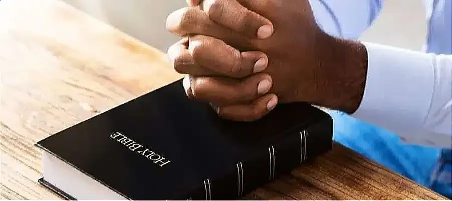 Hands clasped in prayer over a Holy Bible on a wooden table, symbolizing achieving reconciliation with God.