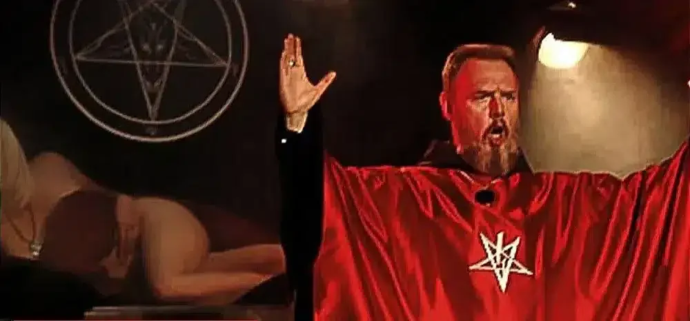 A man in a red robe raises his arms before a pentagram backdrop, exuding power and fervor as part of satan's tactics.